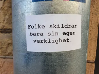A white sticker with black text on a metal surface. The text is in Norwegian and translates to "People only describe their own reality."