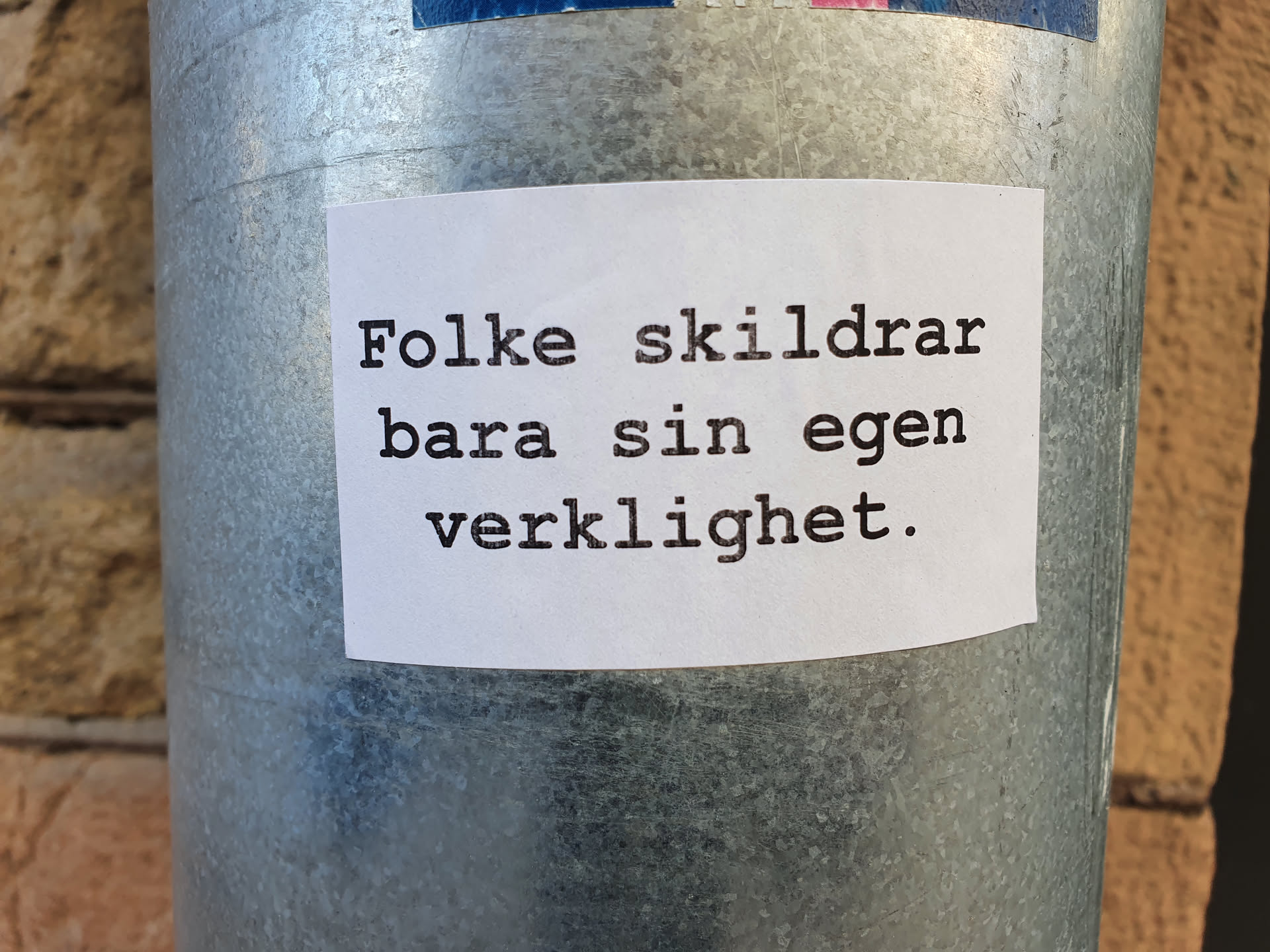 A white sticker with black text on a metal surface. The text is in Norwegian and translates to "People only describe their own reality."