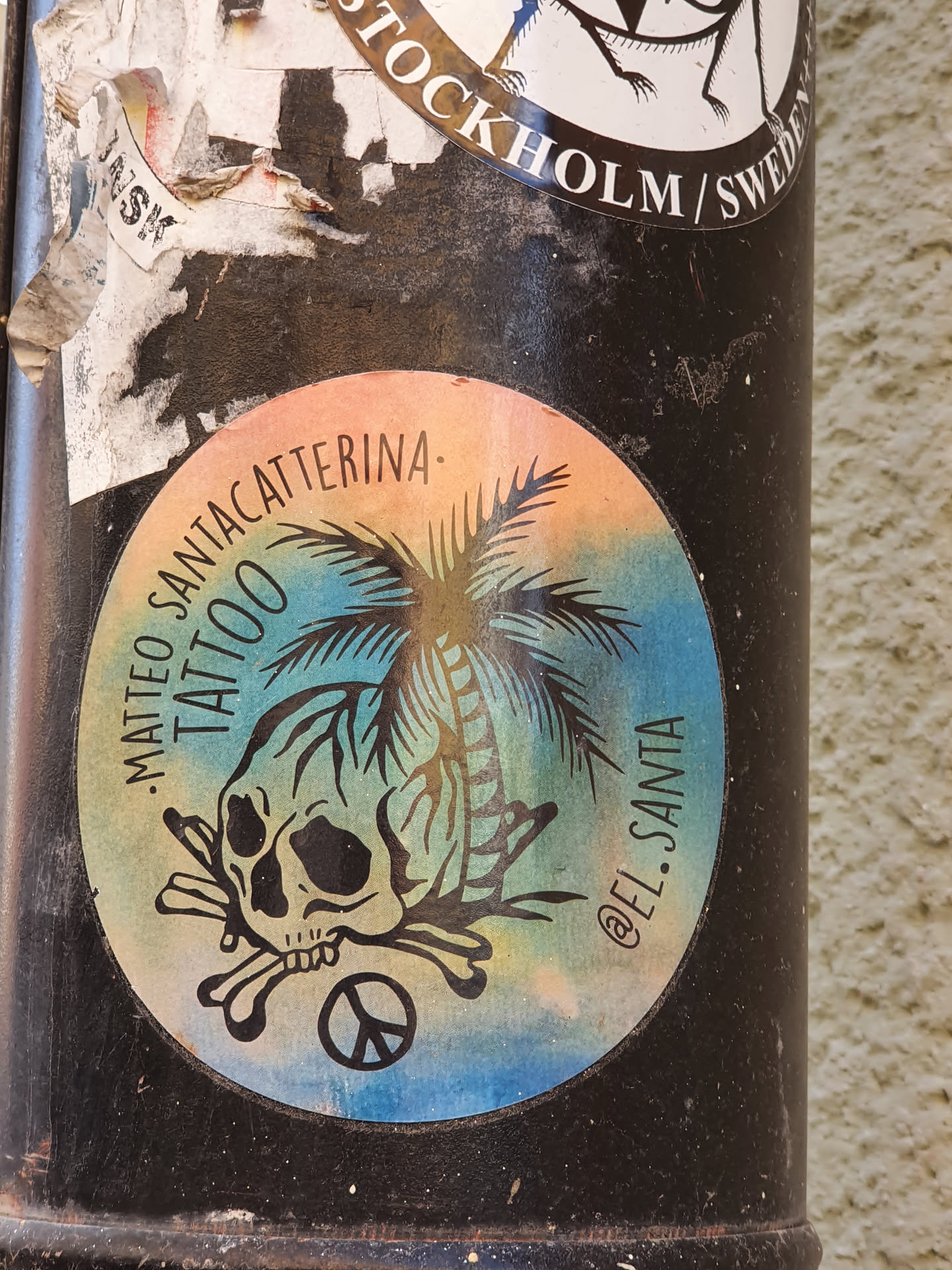 Round sticker with a colorful design featuring a skull with crossbones, a palm tree, and a peace symbol. The text 'MATEO SANTACATERINA TATTOO' and '@EL.SANTA' are visible on the sticker. The background is a gradient of blue and orange colors.