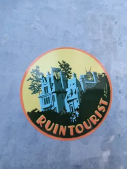 A round sticker with an illustration of a dilapidated building nestled among trees. The words "Ruin Tourist" are written around the central image. The sticker has a yellow background, dark green border, and orange outline.