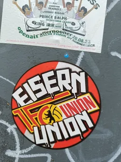 A circular sticker featuring the Eisern Union logo, predominantly red and white with black text.  The logo includes a stylized bear emblem. The sticker appears to be affixed to a gray surface.