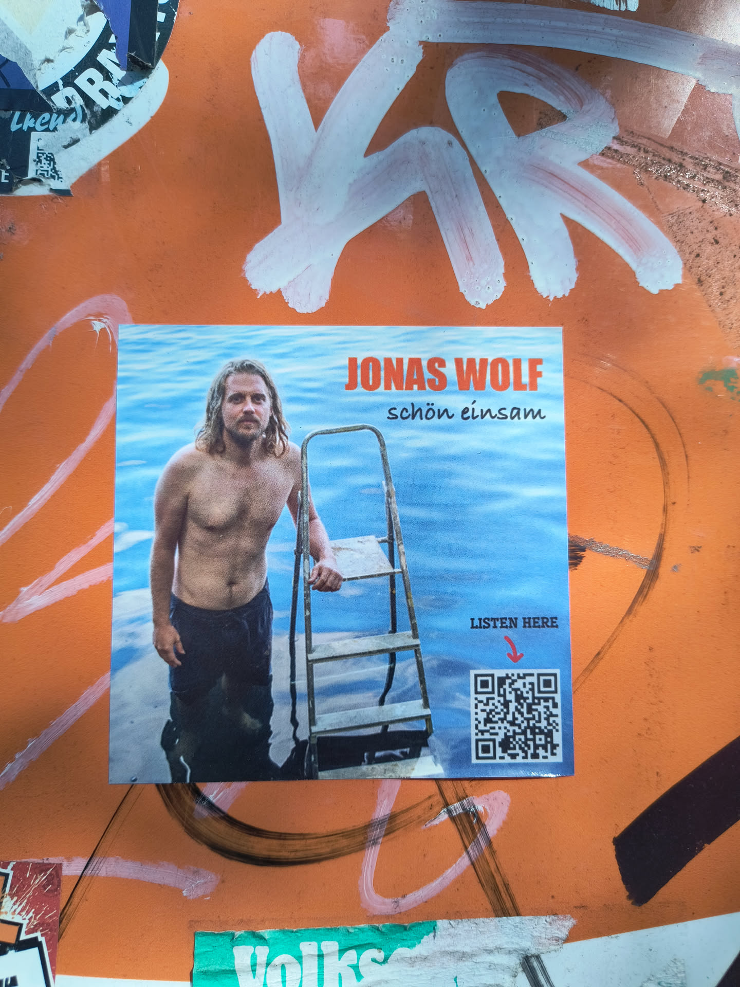 The sticker features a square, full-color photograph of a man with long, light brown hair and a beard, standing shirtless in calm, light blue water. He is wearing dark shorts or trousers. A grey metal ladder with visible wooden steps is positioned to his right, partially submerged in the water. The background is entirely the serene blue water. In the upper right corner, the name "JONAS WOLF" is displayed in bold orange capital letters, with "schön einsam" in smaller orange cursive text directly below it. The lower right corner contains the words "LISTEN HERE" in black, followed by a small red downward-pointing arrow. Below this text, a prominent black and white QR code is present.