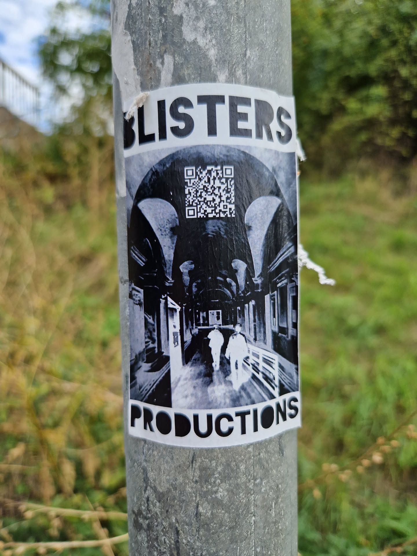 A black and white sticker featuring the text 'Blisters Productions' and a QR code. The main image depicts two figures walking down a dimly lit, arched alleyway.