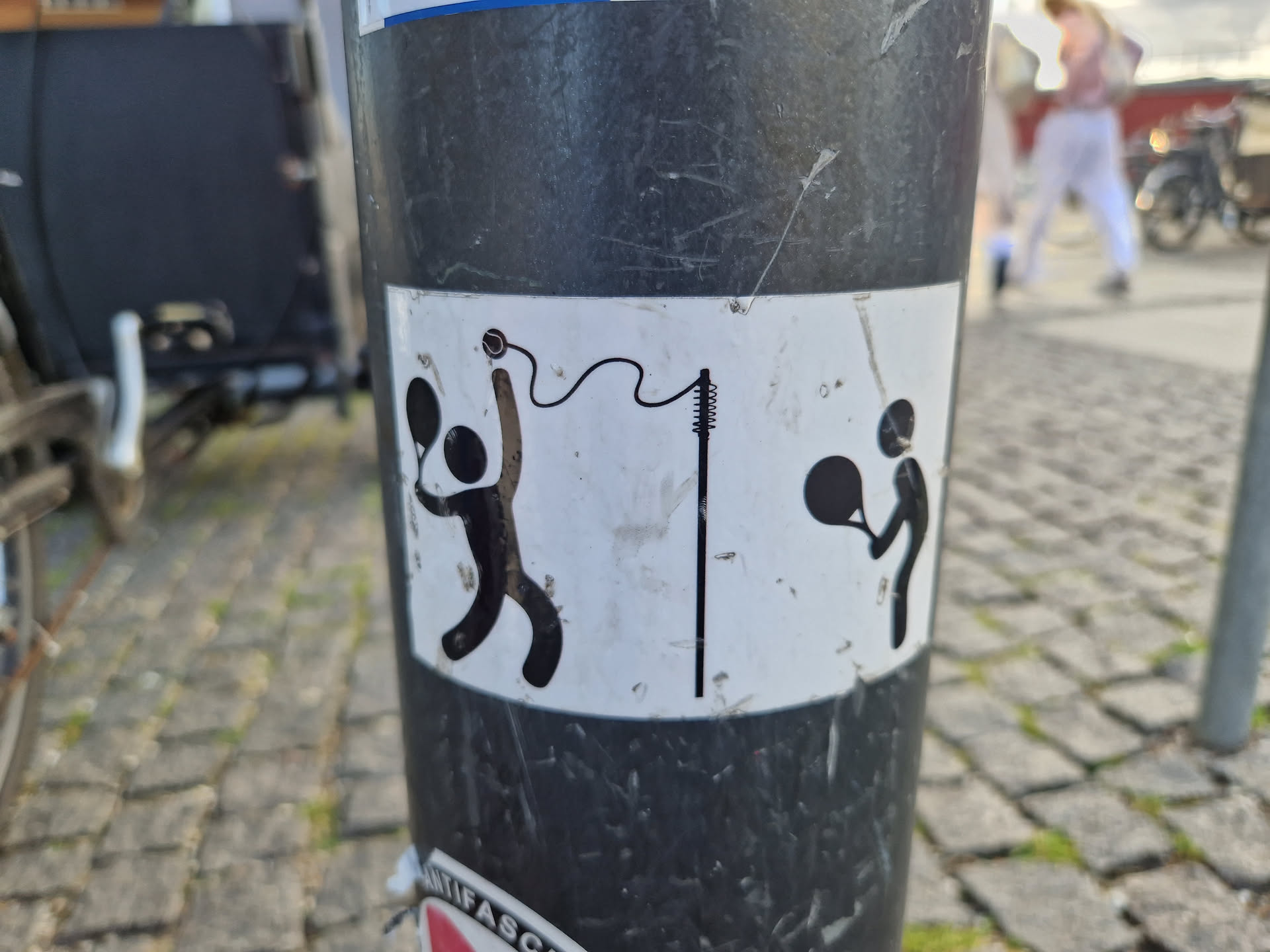 A sticker featuring a minimalist design of two figures playing tennis. One is serving, the other receiving. A tennis net is also depicted. The style is simple and playful.