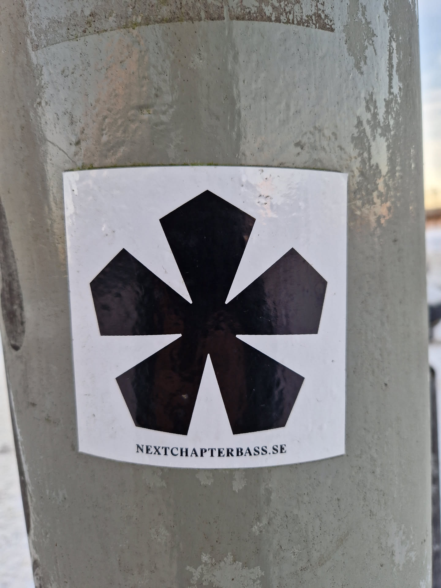 A white square sticker featuring a black, stylized flower or starburst graphic.  Text at the bottom reads 'NEXTCHAPTERBASS.SE'.