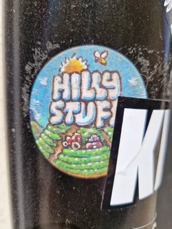 A round sticker with a blue background depicts a hilly landscape with a sun, a bee, and cartoonish farm elements. The text 'Hilly Stuff' is prominently displayed in stylized lettering. The sticker appears to be affixed to a dark-colored surface.
