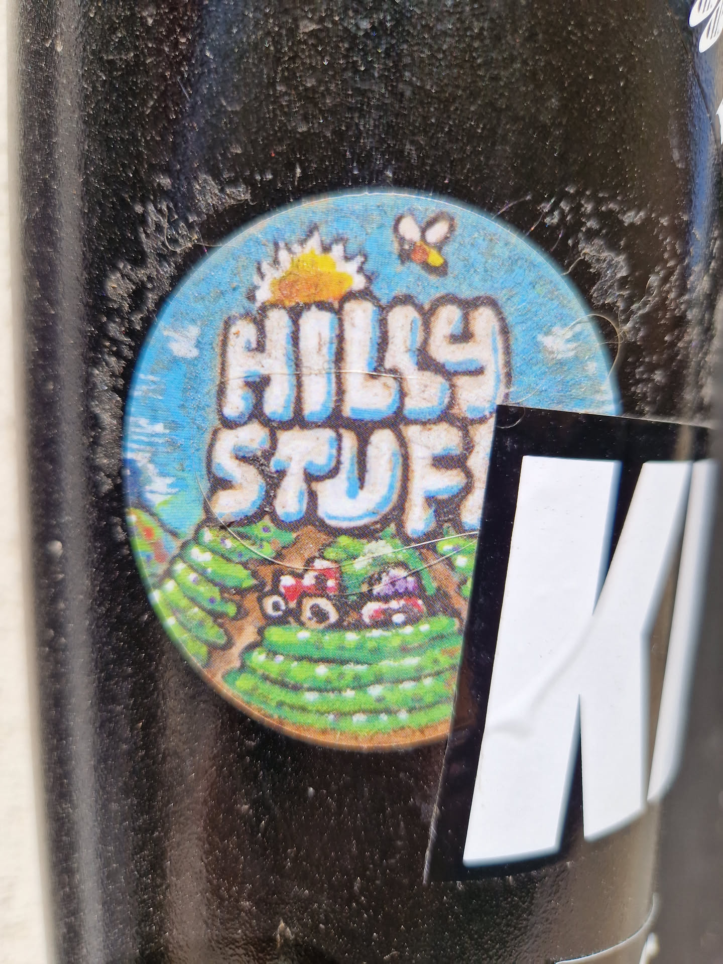 A round sticker with a blue background depicts a hilly landscape with a sun, a bee, and cartoonish farm elements. The text 'Hilly Stuff' is prominently displayed in stylized lettering. The sticker appears to be affixed to a dark-colored surface.