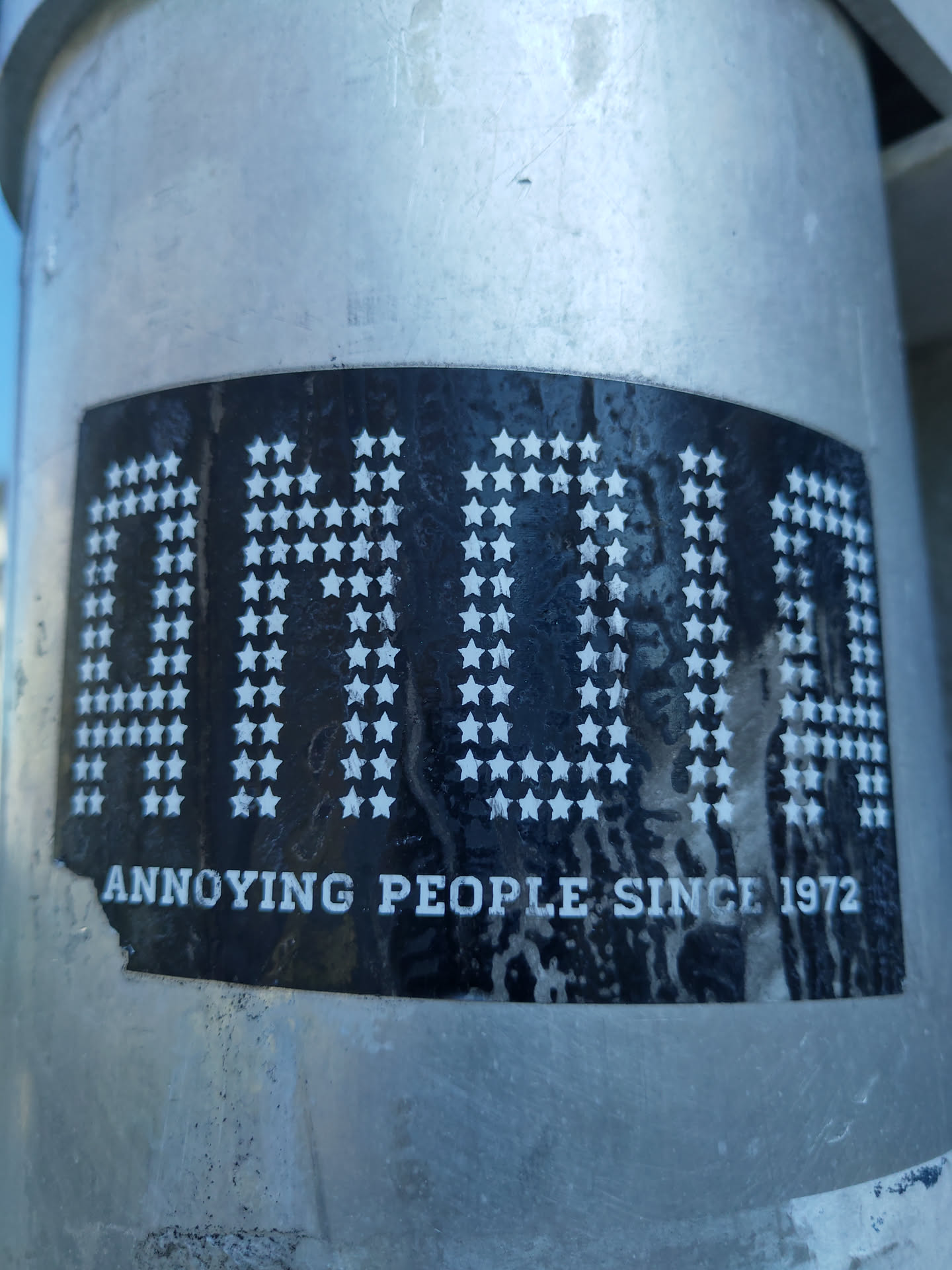 A black and white sticker with the text "AH-HA" in a unique font made of stars and the words "Annoying People Since 1972" underneath. The sticker appears to be affixed to a metallic surface.