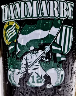 A sticker featuring the Hammarby Football Club logo. It includes the club's name, a figure with a pole, and the number 12. The sticker is wet and has water droplets on it.