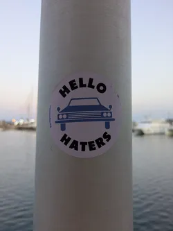 This is a round, light pink sticker with a dark blue car graphic in the center. The words "HELLO" are curved above the car and "HATERS" is curved below it. The sticker appears on a light post in a marina or harbor setting.