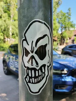A black and white sticker of a skull with a wide, toothy grin affixed to a metal post. The skull is drawn in a simple, almost cartoonish style. 