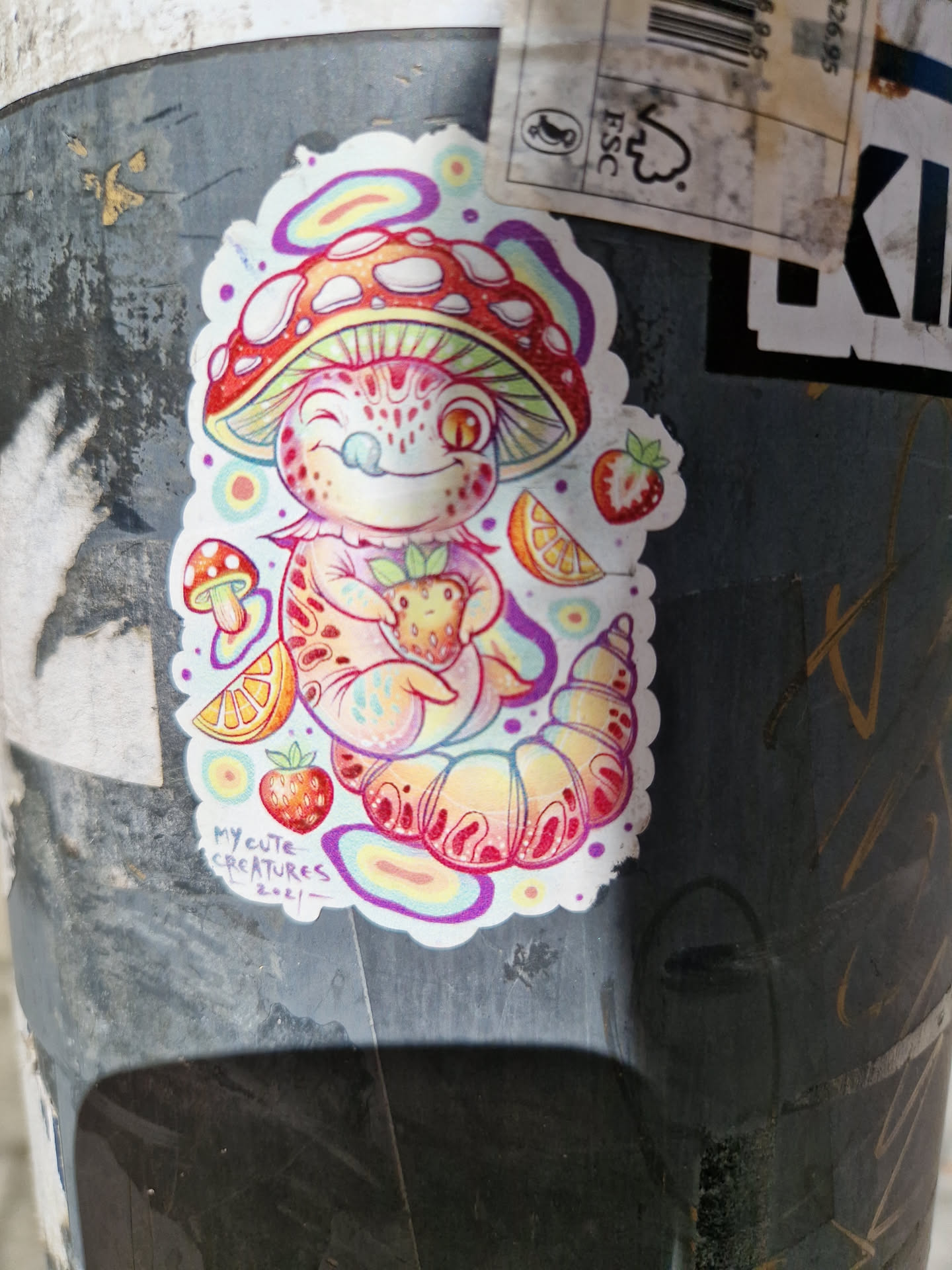A cute sticker featuring a cartoon creature under a mushroom, surrounded by strawberries and orange slices. The creature appears happy and is holding a small strawberry. The artwork style is vibrant and colorful.