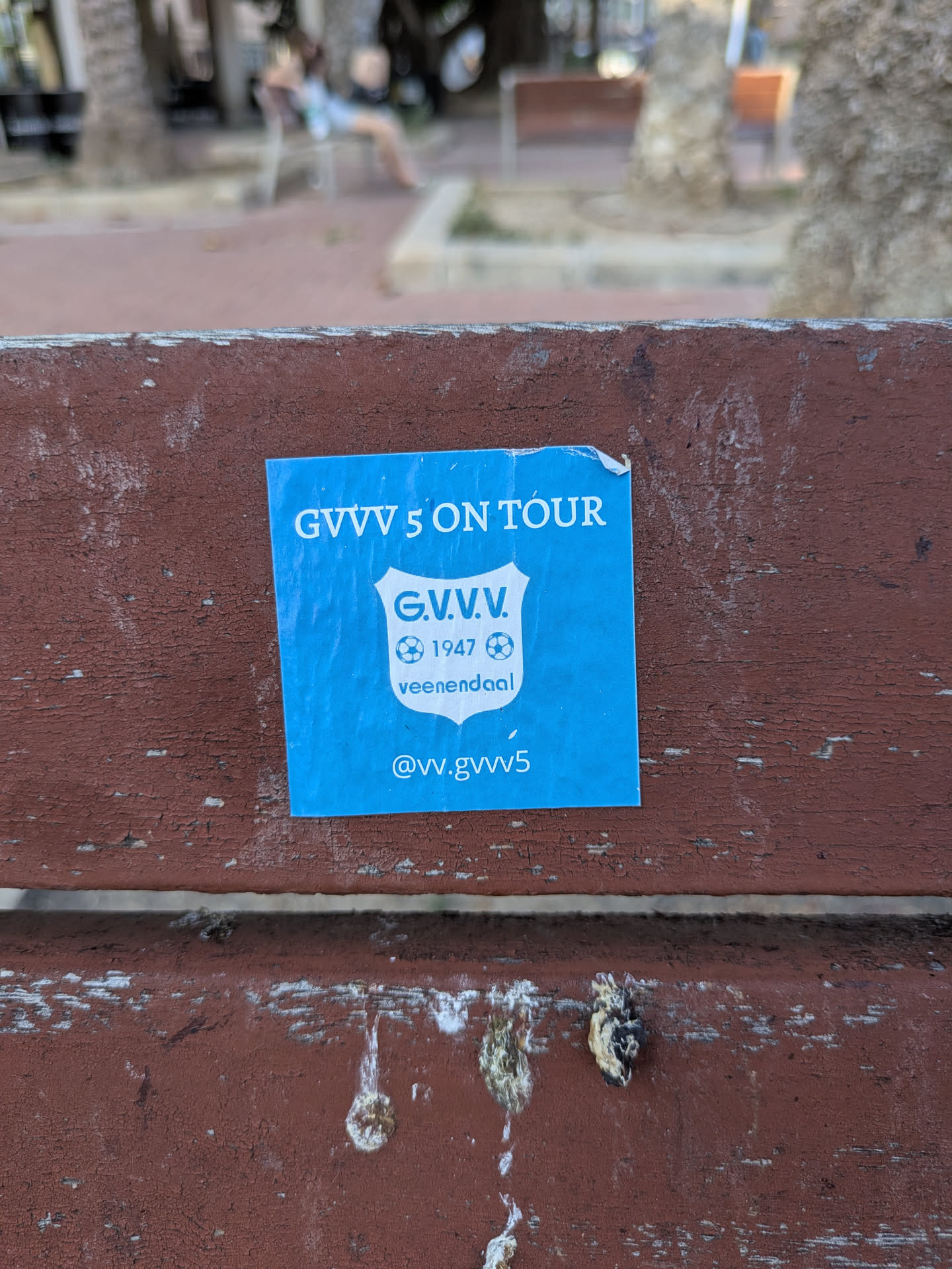 A blue square sticker is stuck on a wooden bench. The sticker features the text "G.V.V.V. 5 ON TOUR" at the top, the team's crest in the middle, and the Instagram handle @vv.gvvv5 at the bottom. The crest includes the text "G.V.V.V." "1947" and "veenendaal".