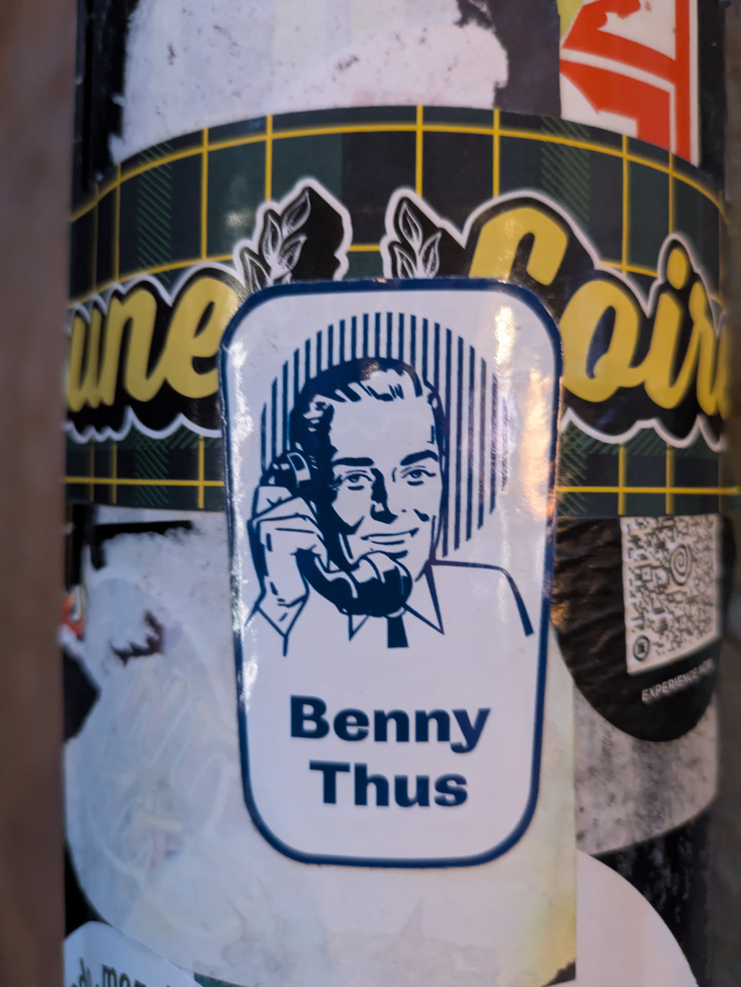 The sticker features a stylized blue and white illustration of a man holding a telephone to his ear. He has dark hair, a smiling expression, and is wearing a collared shirt and tie. The background behind his head consists of vertical blue lines. The overall shape of the sticker is a rounded rectangle with a thick blue border. Below the illustration, the name 'Benny Thus' is printed in bold blue capital letters. The sticker is affixed to a surface with a checkered pattern (dark green and yellow) in the background, and other stickers are partially visible around it.