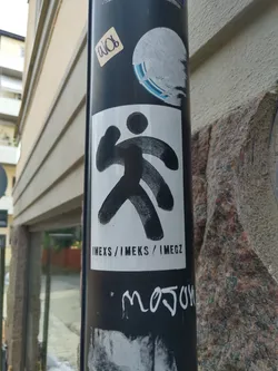 A white sticker with a black abstract figure and the text IMEXS / IMEKS / IMEGZ. The sticker is on a pole with other stickers and graffiti.