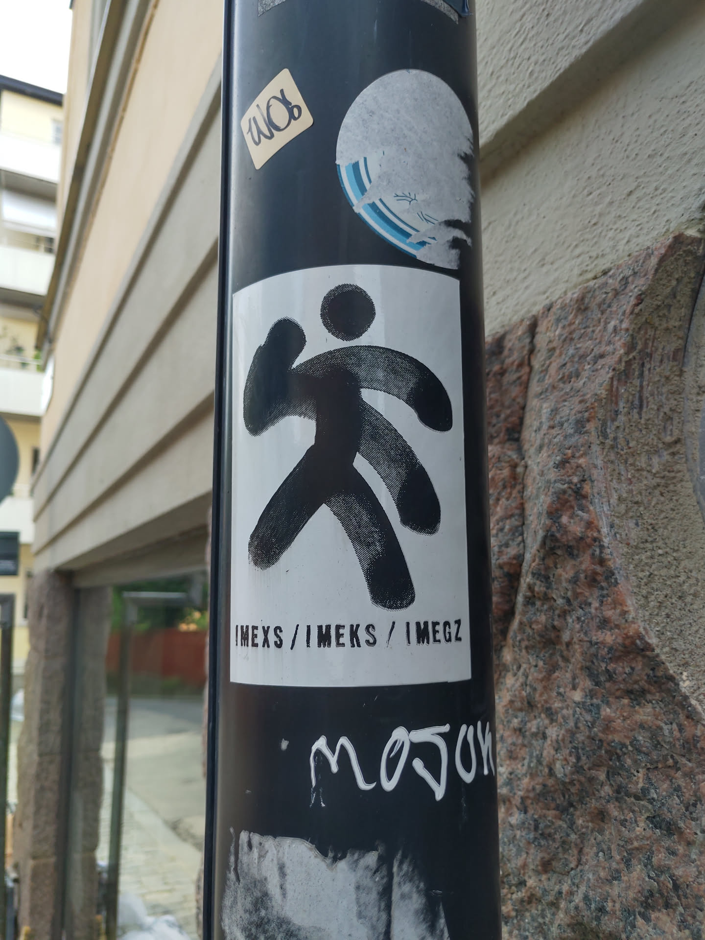 A white sticker with a black abstract figure and the text IMEXS / IMEKS / IMEGZ. The sticker is on a pole with other stickers and graffiti.