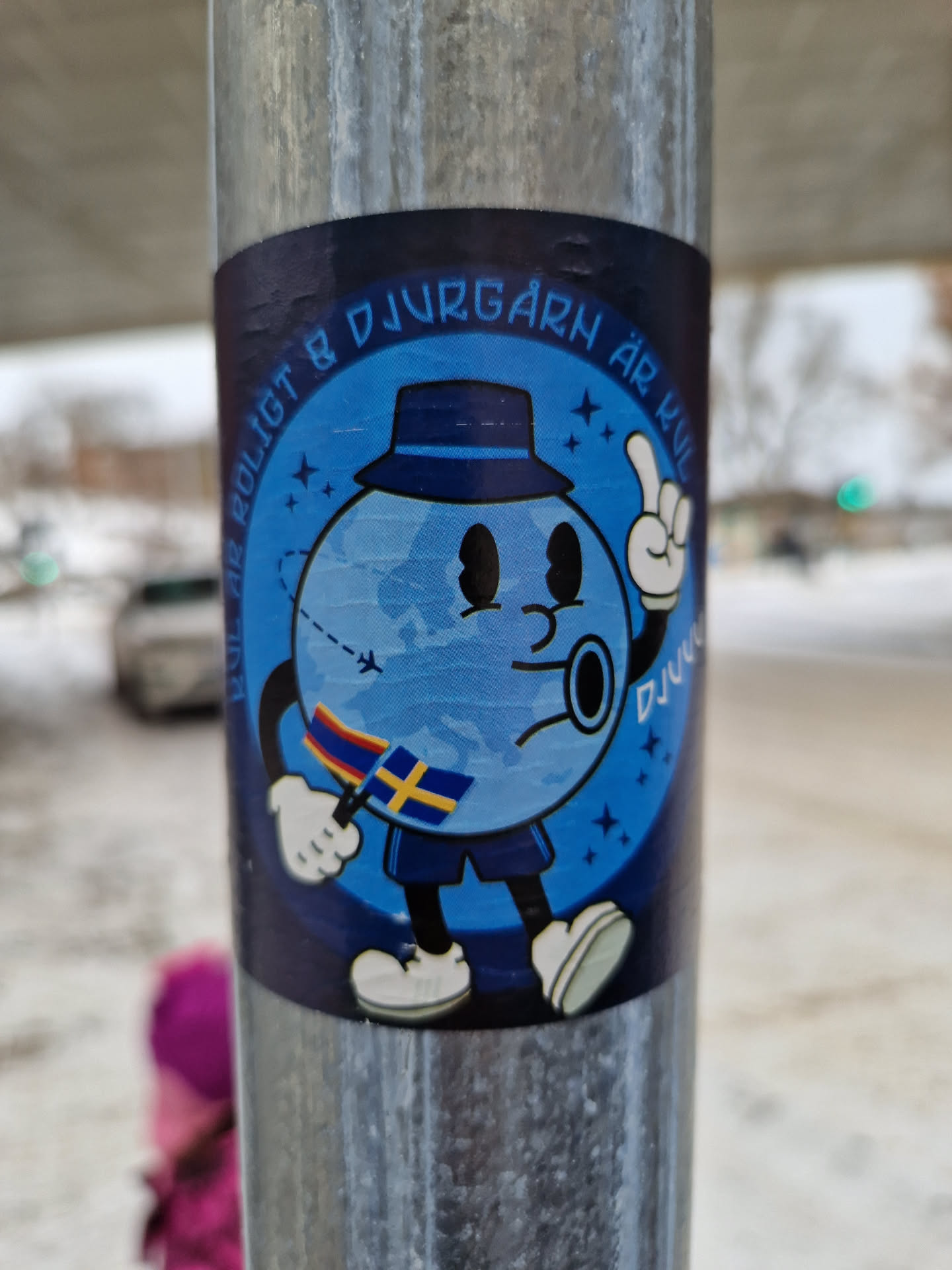 A sticker featuring a cartoon character resembling planet Earth, wearing a hat and holding a Swedish flag. The character is styled in a retro manner, with simple lines and bright colors. The background is a dark blue with stars. The text on the sticker says "Det är roligt på Djurgården är kul".
