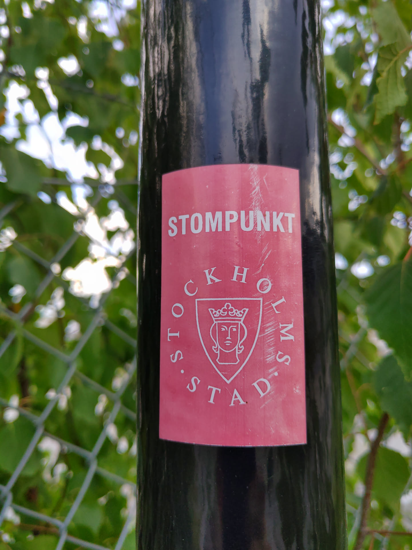 A pink sticker with the text "STOMPUNKT" at the top and a circular design featuring a shield with a crowned face and the text "STOCKHOLMS STAD" underneath. The sticker is affixed to a black pole.