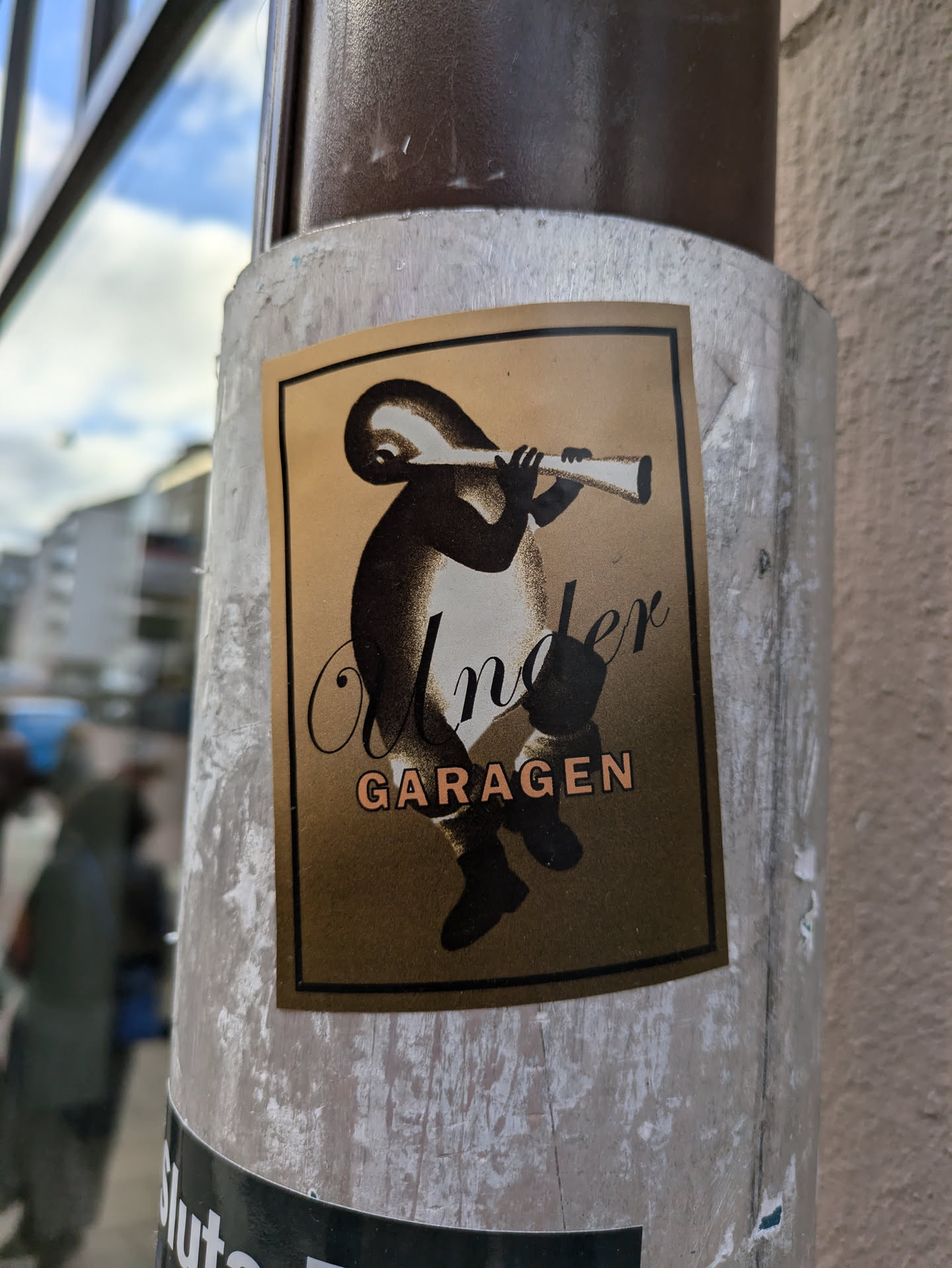 Gold-colored sticker featuring a minimalist illustration of a figure playing a long instrument. The text 'Under Garagen' is prominently displayed.