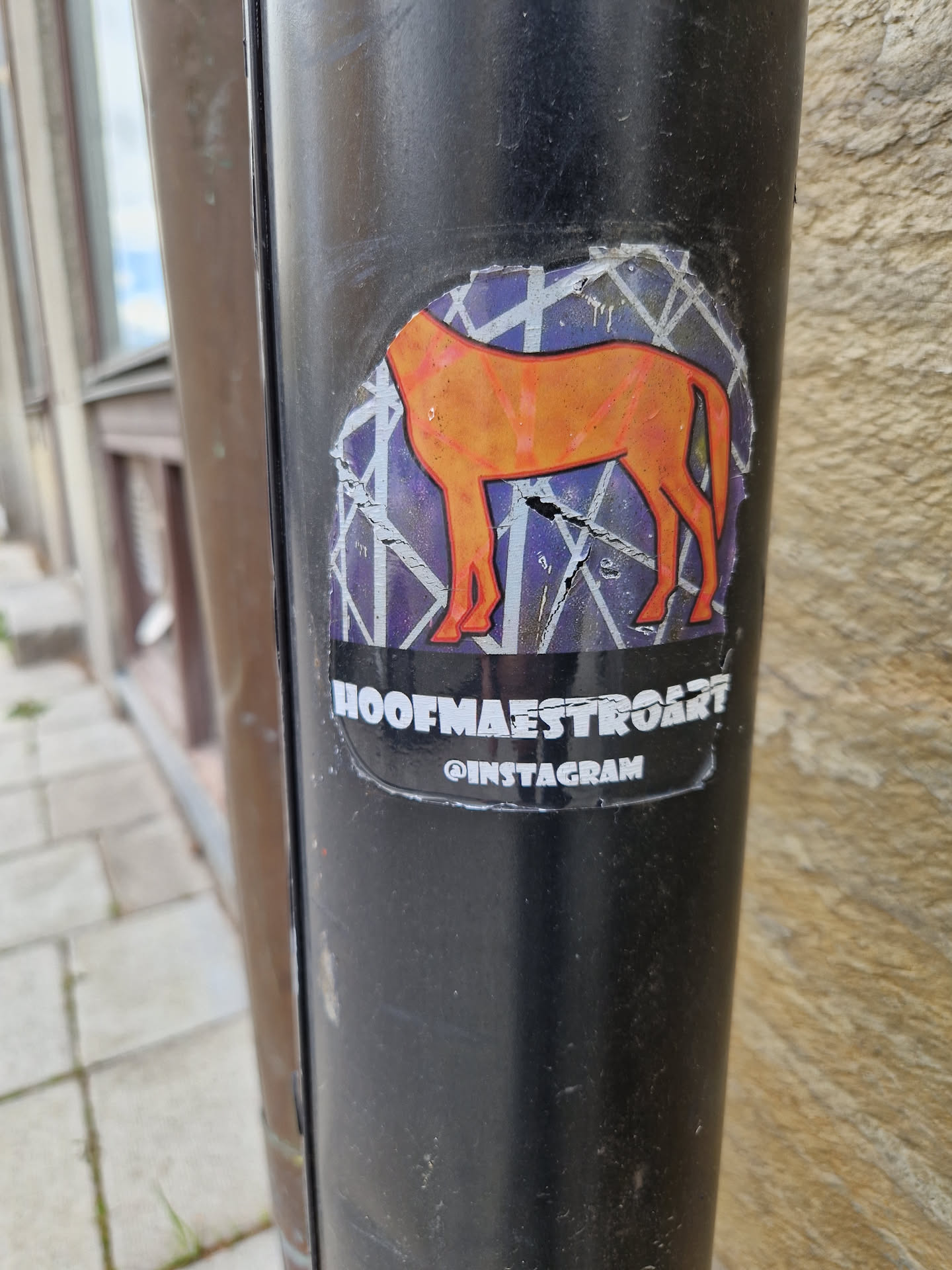 A sticker featuring an orange horse illustration on a geometric background. The sticker is slightly damaged and is affixed to a dark-colored pole.