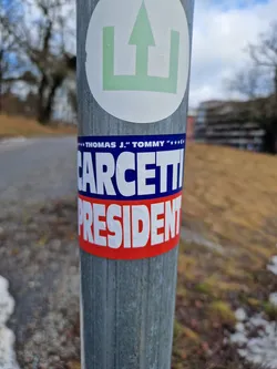 A political campaign sticker for Thomas J. "Tommy" Carcetti for President. The sticker is blue, white, and red and is affixed to a metal pole.