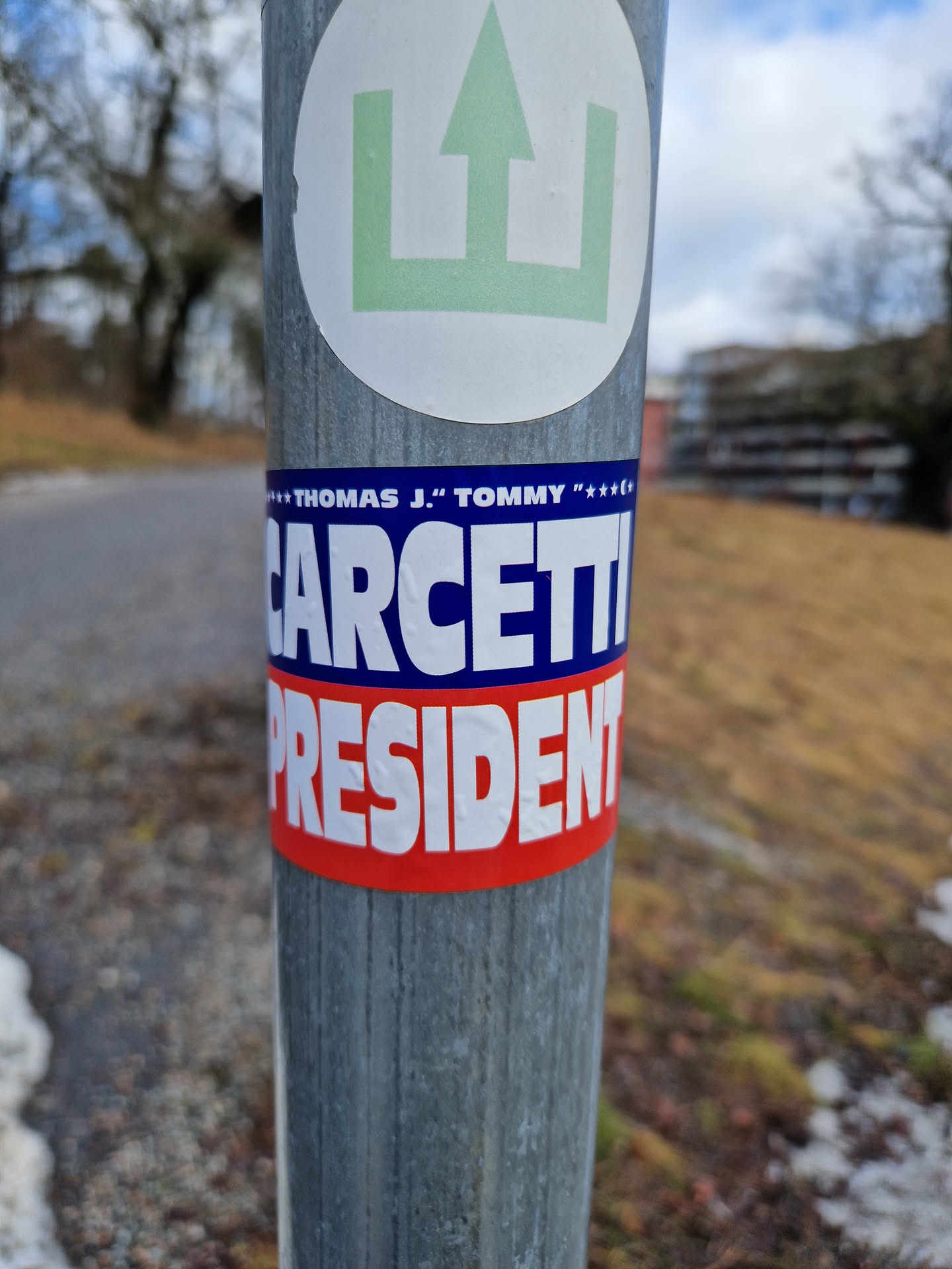 A political campaign sticker for Thomas J. "Tommy" Carcetti for President. The sticker is blue, white, and red and is affixed to a metal pole.