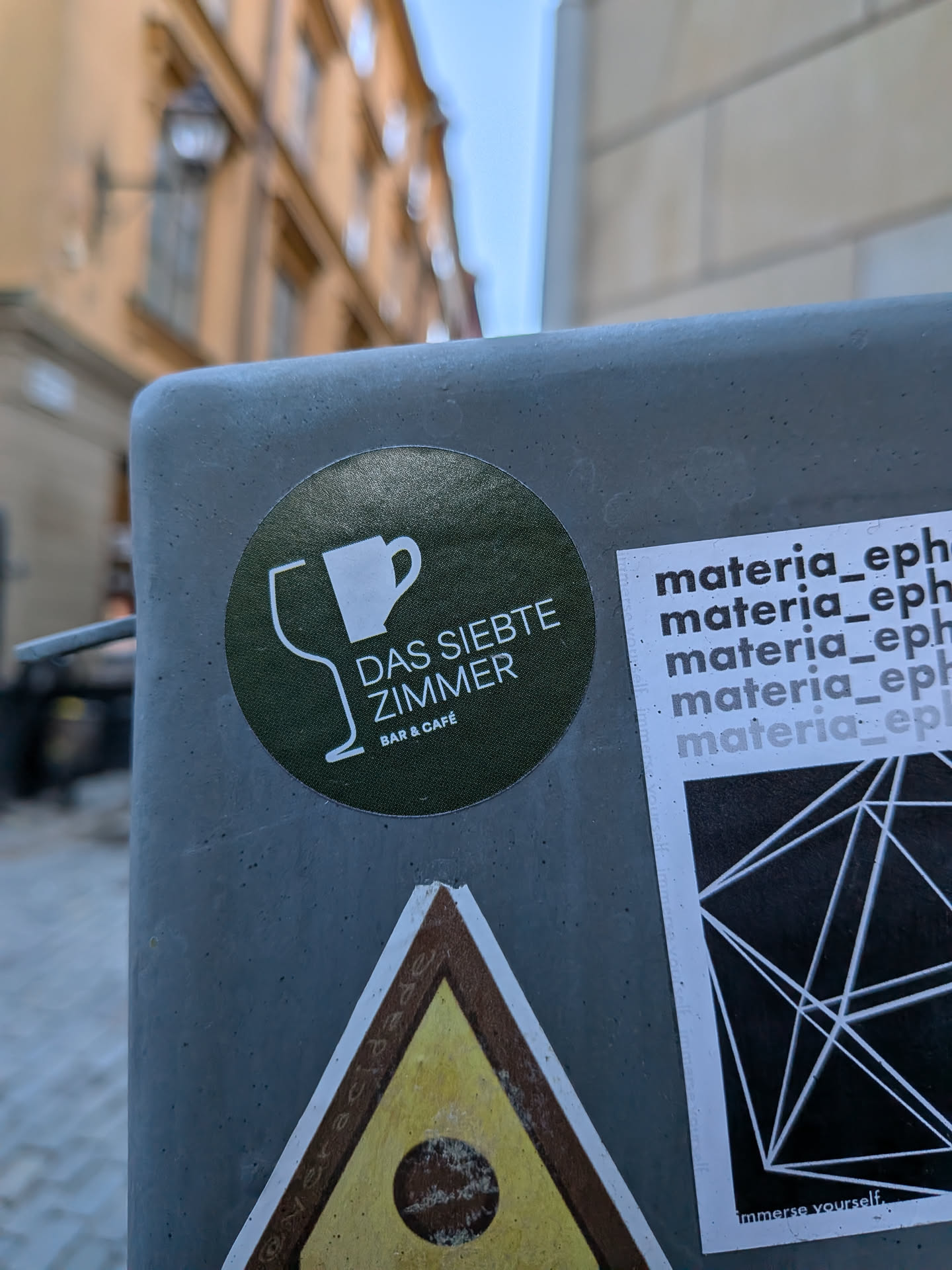 A circular sticker with a dark olive green, slightly textured background. It features two white line-art icons: a stylized wine glass on the left and a coffee mug on the right. To the right of these icons, the text 'DAS SIEBTE ZIMMER' is stacked vertically in white, sans-serif capital letters. Below this, in smaller white text, are the words 'BAR & CAFÉ'. The overall design is clean and minimalist.
