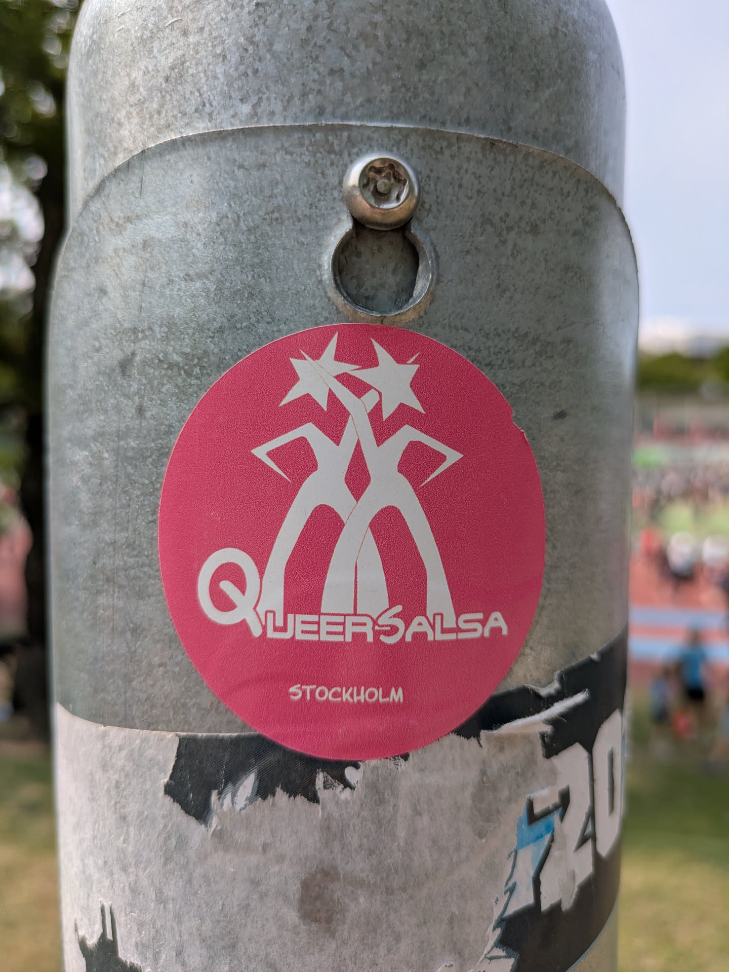 A round, pink sticker is affixed to a metal pole. The sticker has the words 'Queer Salsa' stacked vertically on the left side. To the right of the text are two white figures dancing, their arms reaching upwards, with starbursts at the top of each figure. Below the text and figures, the word 'Stockholm' is printed in smaller letters.