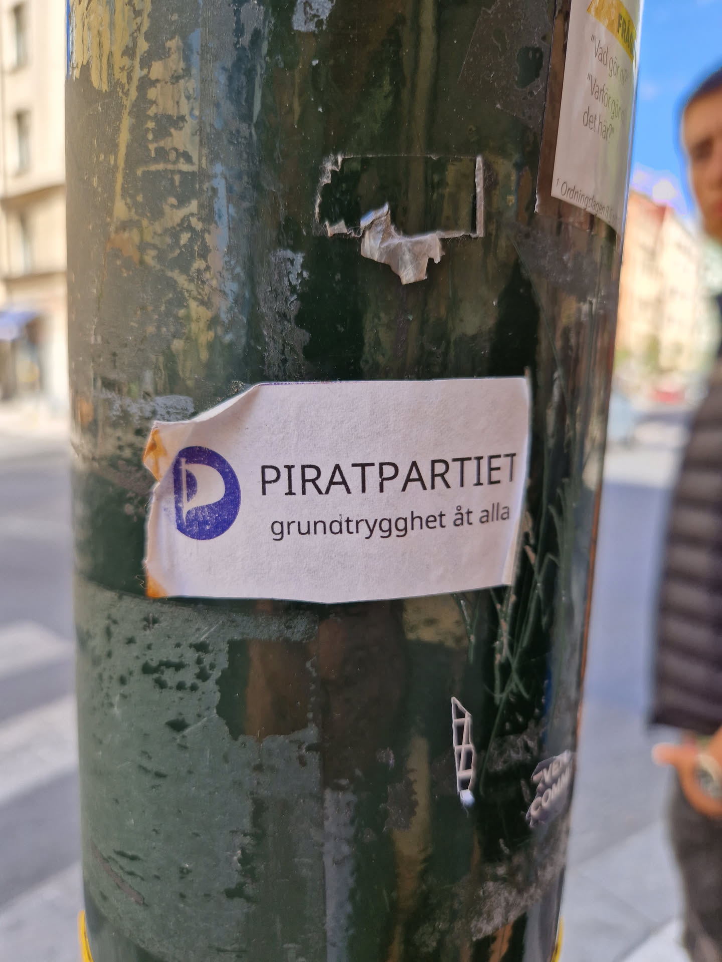 A sticker for the Swedish Pirate Party (Piratpartiet) affixed to a pole. The sticker features the party's logo and the slogan "grundtrygghet åt alla" which translates to "basic security for all".