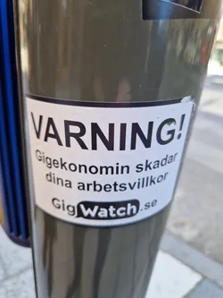 A sticker warning about the negative effects of the gig economy on working conditions.  It features the text "VARNING! Gig ekonomin skadar dina arbetsvillkor" and the logo and website address for GigWatch.se