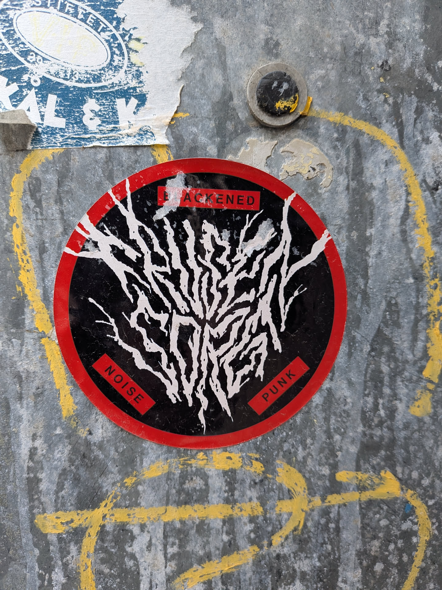 A circular sticker with a bold red outer ring enclosing a black inner circle. The central design features an intricate, chaotic pattern of thick white lines, resembling tangled roots, veins, or a lightning strike, spreading across the black background. Three small, rectangular red boxes with white text are positioned around the central design: 'BLACKENED' at the top, 'NOISE' at the bottom left, and 'PUNK' at the bottom right. The sticker shows signs of wear and tear, with slight peeling and scuffs, indicating it's adhered to a textured, possibly metallic, surface.