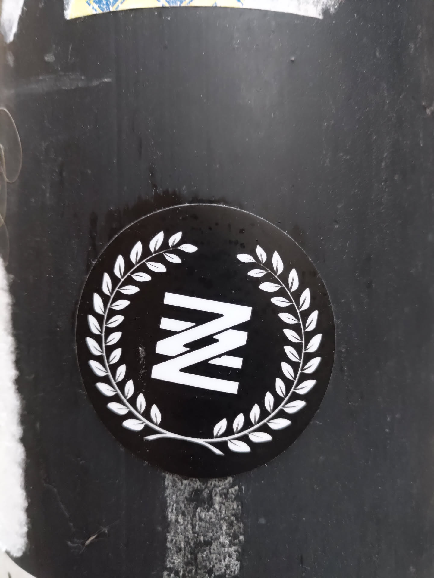 A round black sticker with a white laurel wreath encircling a stylized letter 'N' logo. The logo consists of three vertical, parallel lines forming the shape of a letter 'N'.