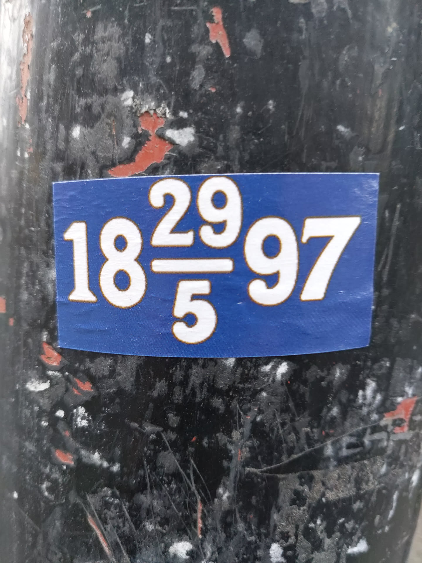 A blue sticker with white numbers, reading "18 29/5 97", is affixed to a weathered, dark-colored pole. The numbers are large and bold, suggesting a possible code or reference. The sticker is rectangular and shows some minor wear from its placement outdoors.
