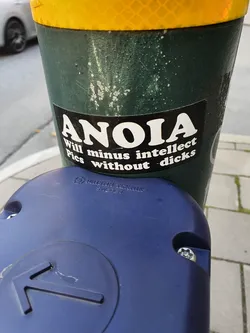 A black and white sticker with the word ANOIA and a phrase that says "Will minus intellect. Pics without - dicks". It's stuck on a green pole.