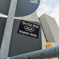 A black and white sticker comparing the Olympic games of Berlin in 1936 and Beijing in 2022. The sticker features the Olympic rings and the years of the respective games.