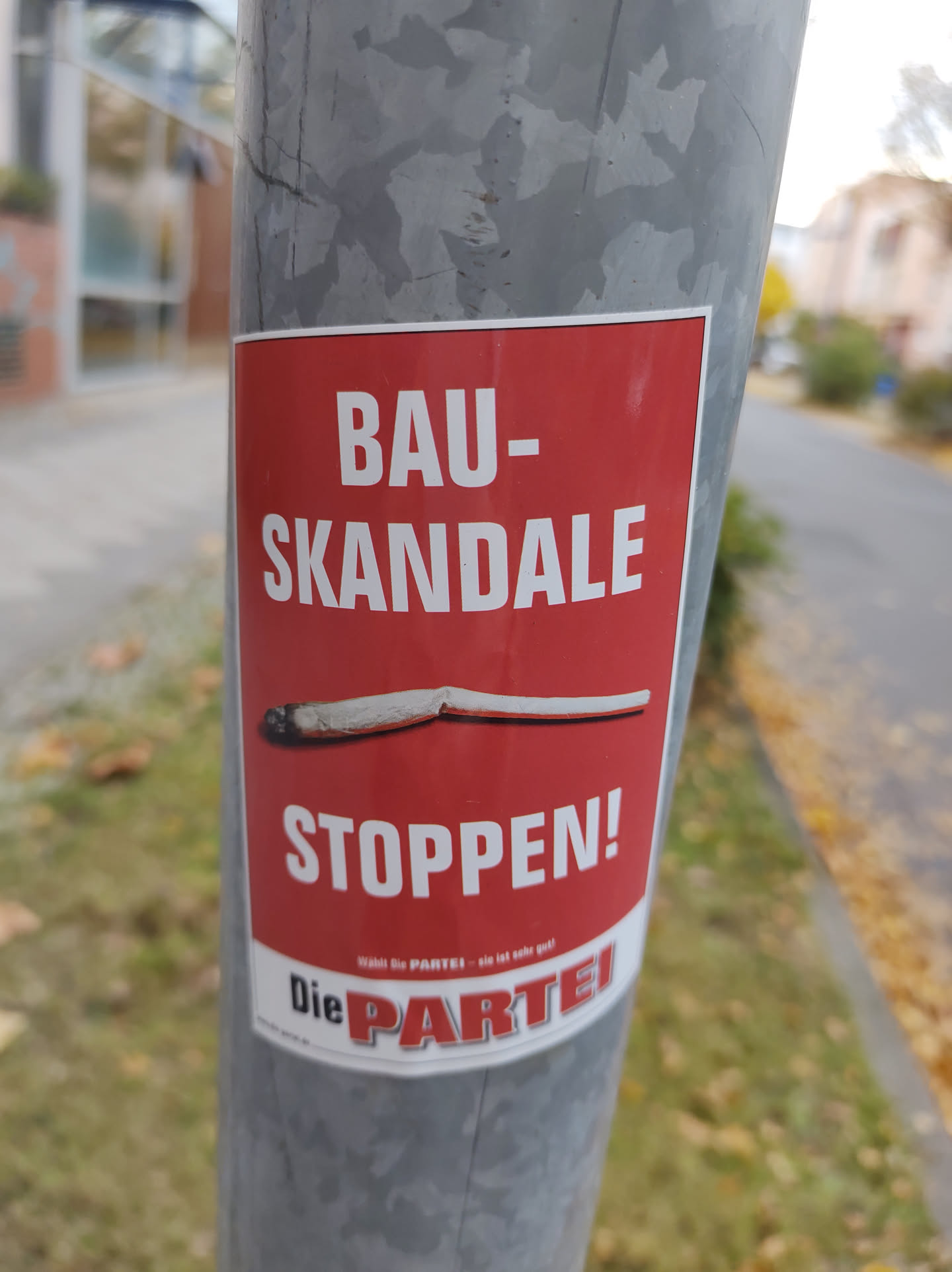 A red sticker with white text on a gray pole.  The text translates to "Construction Scandals - Stop!" with the logo of "Die Partei" (The Party) at the bottom. A broken cigarette is depicted on the sticker.