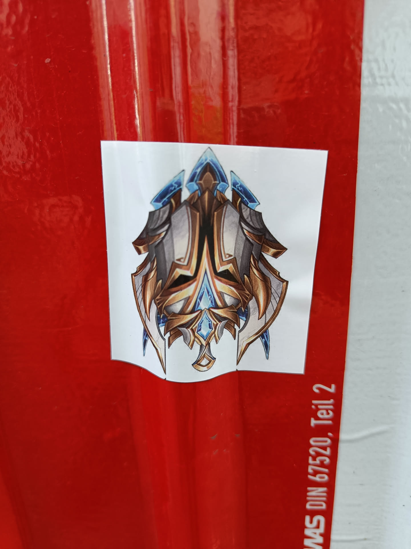 A sticker featuring a detailed drawing of a golden and blue emblem with a sharp, angular design. The emblem is symmetrical and features intricate details.  It's affixed to a red surface.