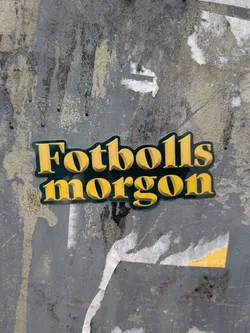 A die-cut sticker featuring the Swedish word 'Fotbollsmorgon' (Football Morning) in two lines. The text is rendered in a bold, retro-style serif font with a prominent 3D effect, appearing in a vibrant golden yellow color. The letters are outlined and backed by a dark forest green, creating a strong contrast and a classic, vintage aesthetic. The sticker is applied to a weathered, grey and beige textured surface.