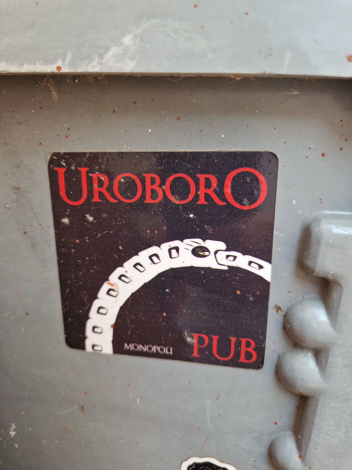 A sticker featuring the word "UROBORO" in red lettering above an image of a snake eating its tail, with "MONOPOLI PUB" written at the bottom. The background is dark with a starry appearance.