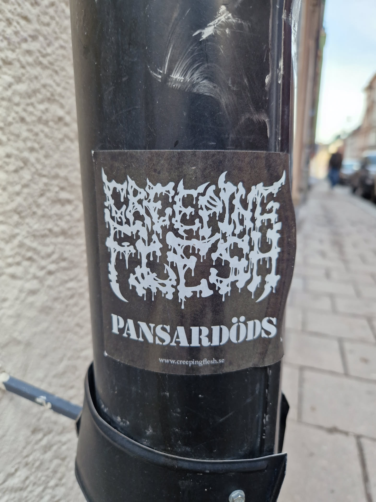 A black and white sticker with a band logo. The logo is in a dripping style and says "CREEPING FLESH" in a stylized font. Below the logo is the band's name, "PANSARDÖDS", and their website address, www.creepingflesh.se. The sticker is placed on a black pole.