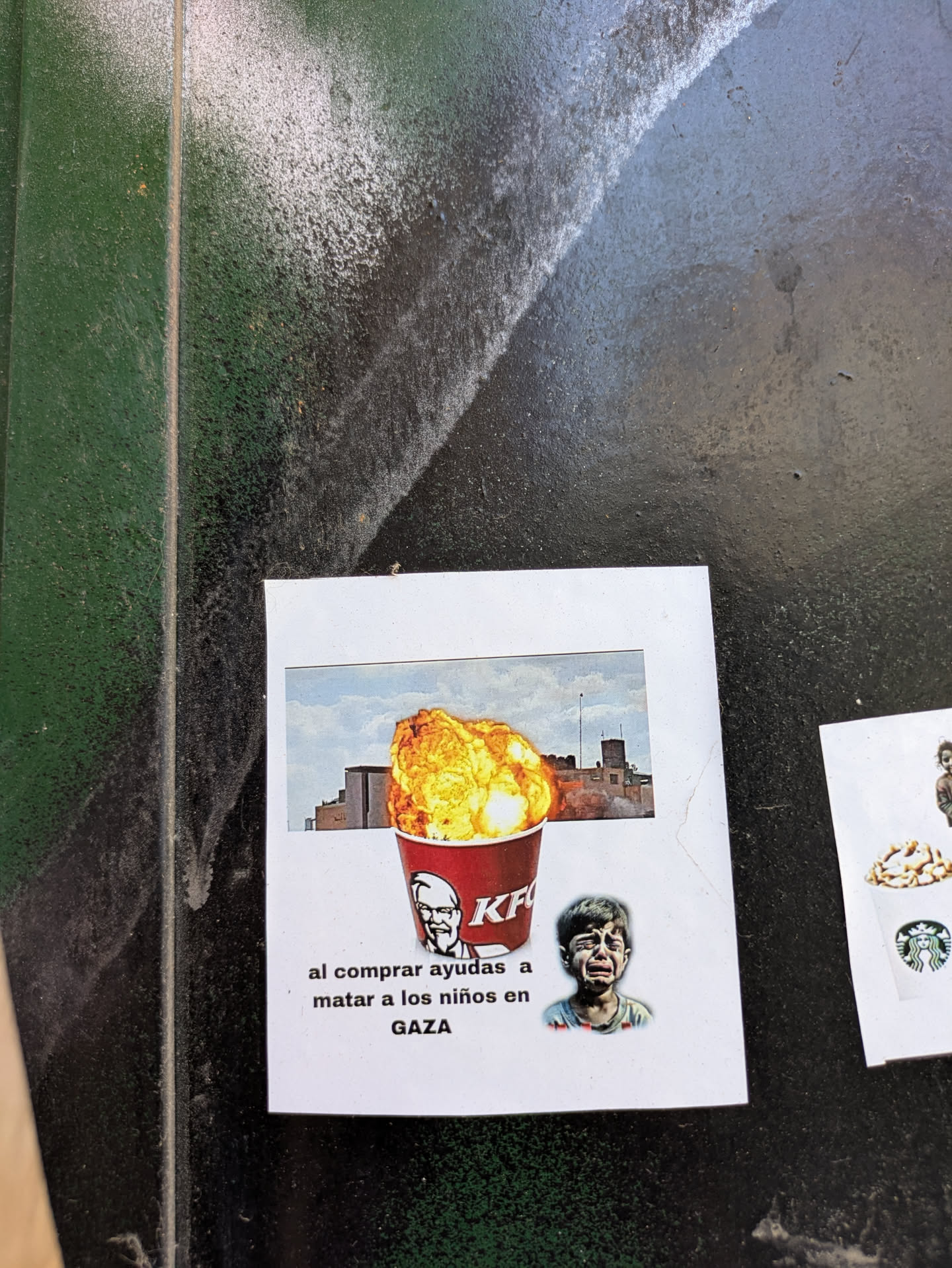 A sticker featuring an altered KFC advertisement. The bucket of chicken has been replaced with an explosion over a building, likely meant to resemble bombings in Gaza. Below this imagery, there's text reading "al comprar ayudas a matar a los niños en GAZA," which translates to "buying helps kill children in GAZA." A crying child's face appears next to the bucket.
