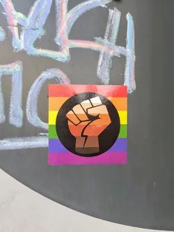 The sticker features a rainbow pride flag with a raised fist in a black circle, showcasing support for LGBT rights and racial equality. The fist is gradient and shades of brown.