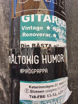 A black sticker with the text "RÅLTOKIG HUMOR" and the Instagram handle @PIROGPAPPA.  The sticker is placed on a pole with other stickers underneath.
