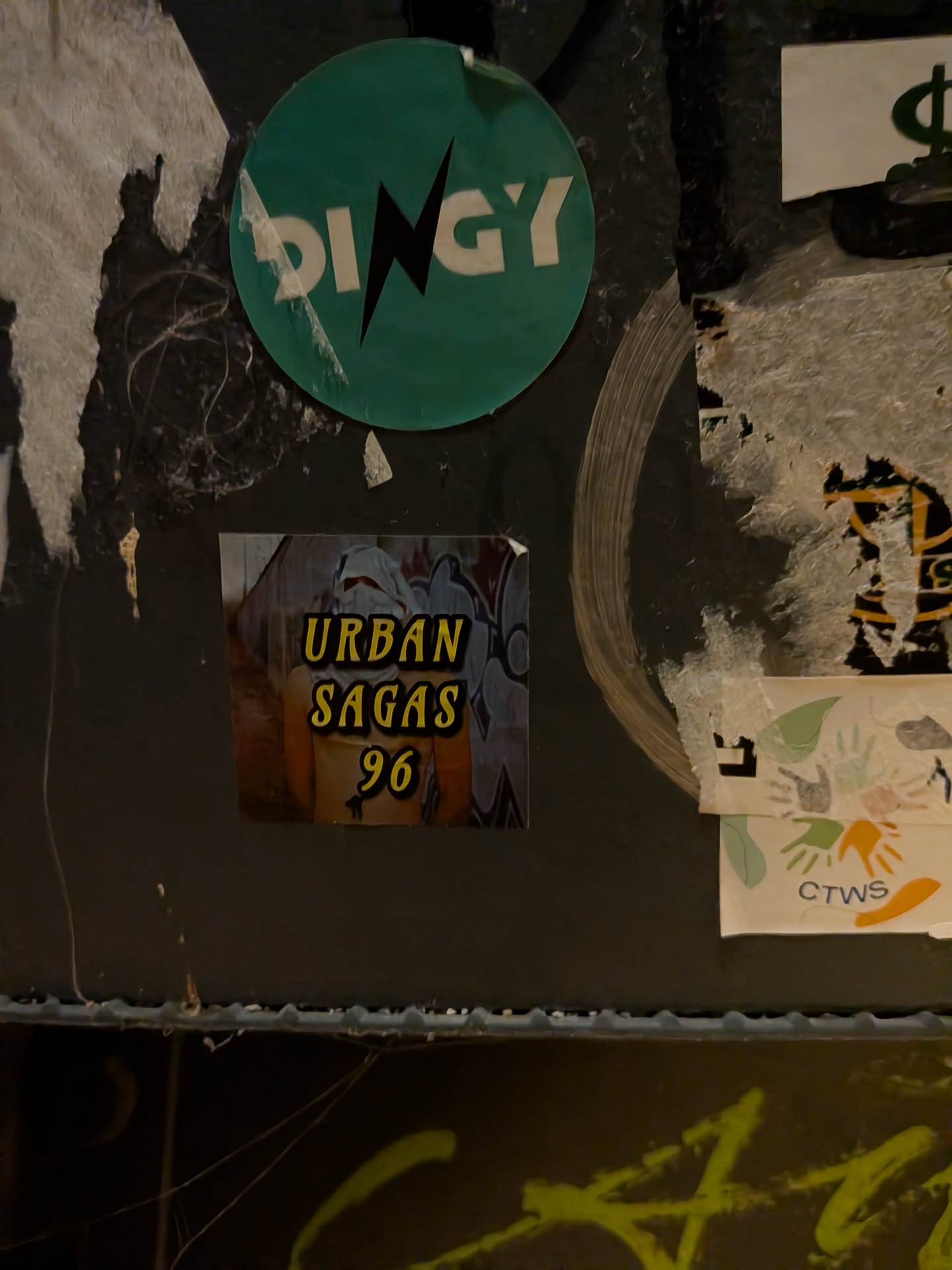 A square sticker affixed to a worn, dark surface. The image on the sticker depicts a masked individual with a white cloth wrapped around their face, standing shirtless in front of a graffiti-covered wall or train. Bold, stylized yellow text with black outlines in the center reads 'URBAN SAGAS 96'. The top-right corner of the sticker is slightly peeled.