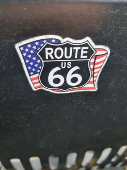A close-up shot of a Route 66 sticker. The sticker features the iconic Route 66 shield with the number 66 in the center.  American flags are depicted on either side of the shield. The sticker is adhered to a dark gray or black surface.