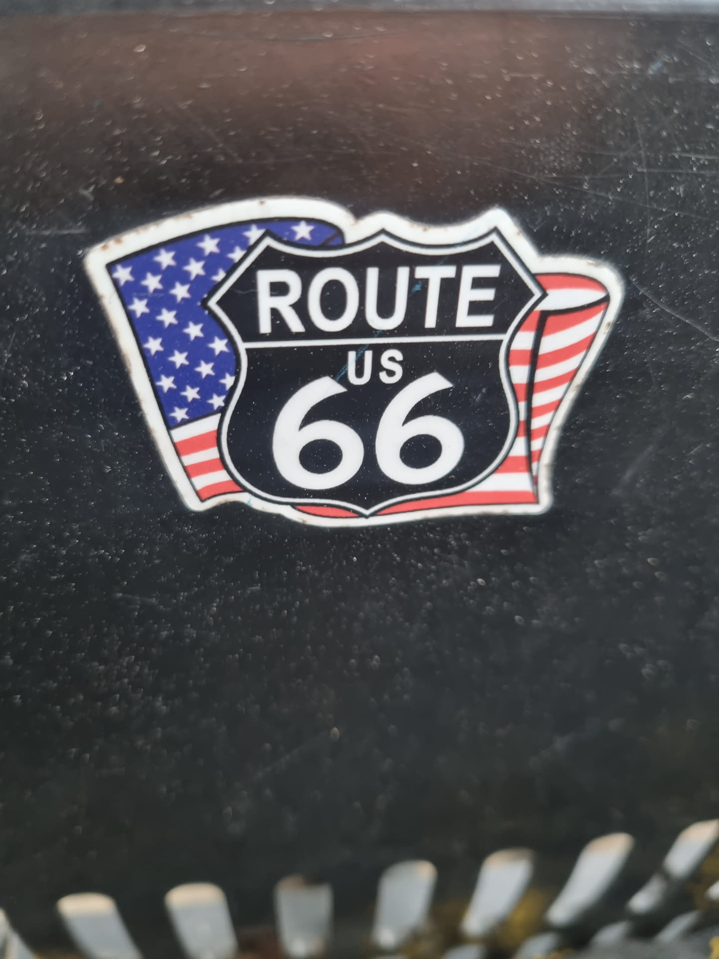 A close-up shot of a Route 66 sticker. The sticker features the iconic Route 66 shield with the number 66 in the center.  American flags are depicted on either side of the shield. The sticker is adhered to a dark gray or black surface.