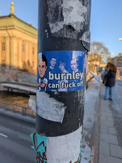 A rectangular sticker with a vibrant blue background, affixed to a metal pole. On the left side, a cartoon caricature of a man with dark hair, glasses, and a light complexion is depicted with his arms crossed. He is wearing a blue and white halved football shirt, likely representing Blackburn Rovers, complete with a small crest. To the right of the cartoon character, against the same blue background, are three real-life young men, possibly footballers or fans, looking forward. Bold white text in a sans-serif font reads 'burnley can fuck off' across the lower right portion of the sticker. The sticker shows some signs of wear and tear, particularly around the edges.