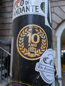 The sticker is circular with a prominent gold/yellow outer border and a black background. In the center, a large, stylized '10' is depicted in gold/yellow, encircled by a gold/yellow laurel wreath. Below the '10', the word 'ANNI' (Italian for years) is written in gold/yellow. Further below, a rectangular emblem features 'ECB' at the top, a silhouette of a she-wolf suckling two infants (a symbol of Rome) in the middle, and 'ETERNAL CITY BREWING' along with 'SINCE 2015' at the bottom, all in gold/yellow on a black background. Curved along the top edge, above the '10' and wreath, are the dates '2015-2025' in gold/yellow. The sticker shows some signs of wear, consistent with outdoor placement.