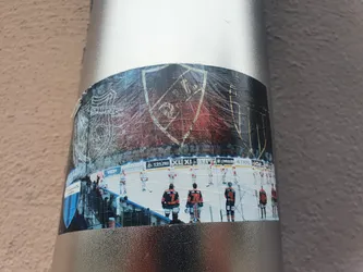 A sticker depicting an ice hockey team. The sticker includes a prominent crest, likely the team's logo, and a photo of players on the ice during a game.  The crest is a shield shape with some text that is difficult to read completely. The image quality of the sticker is slightly degraded, suggesting it may have been exposed to the elements.