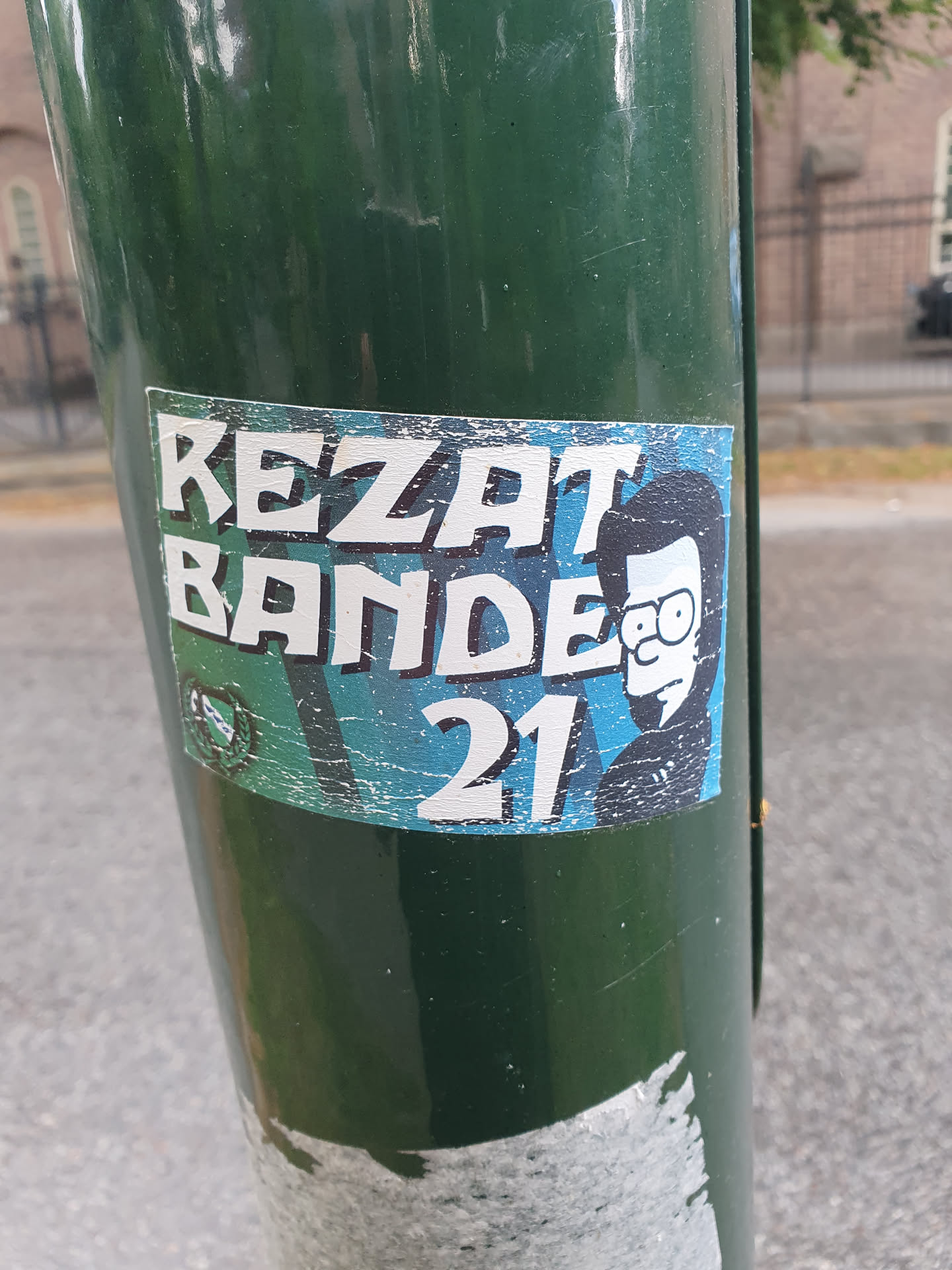 A worn sticker featuring the text "REZAT BANDE 21" and an illustration of a person resembling a cartoon character. The sticker is affixed to a green pole.